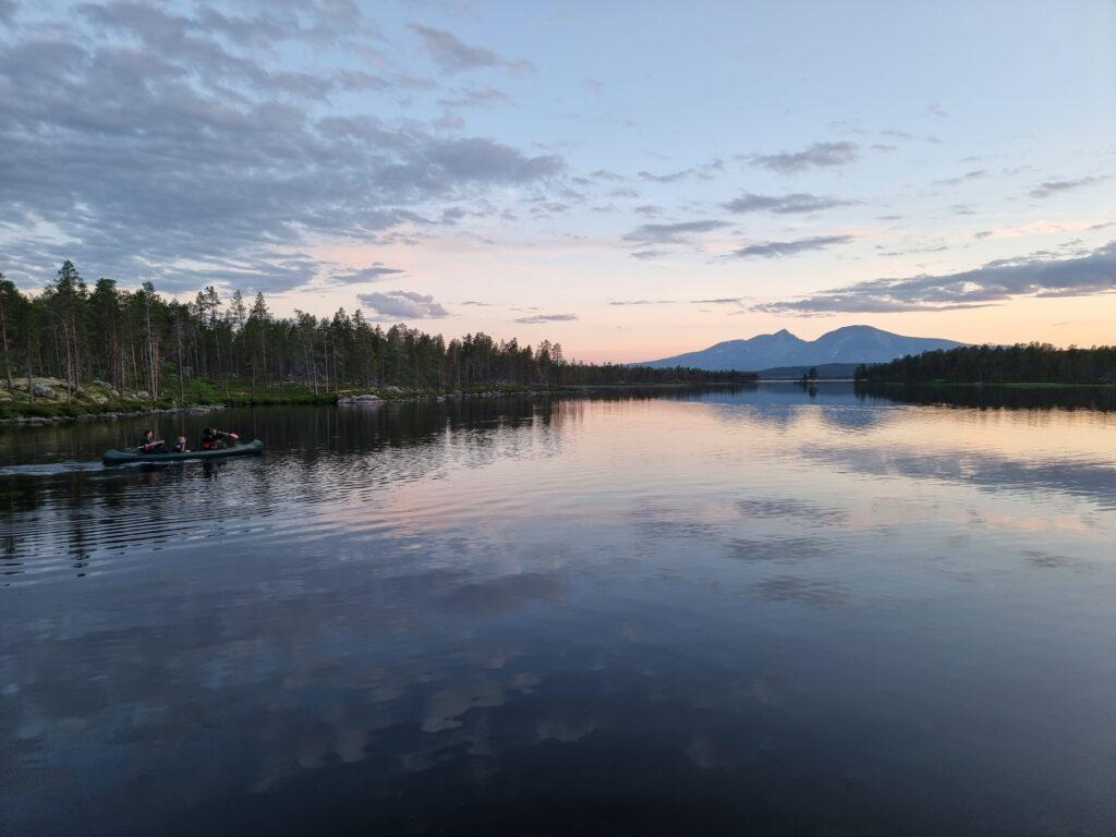 Bilde av en familie ute på kanotur på et blikkstille vann. Familien padler i en blågrønn kano, på blått vann og har en grønn skog i bakgrunnen. Himmelen er blå med solnedgang. De har alle på seg redningsvest. Vi forteller om man må ha våtkort for å padle i kajakk, kano eller packraft? Og hva introkurs og grunnkurs i kajakk og kano er.