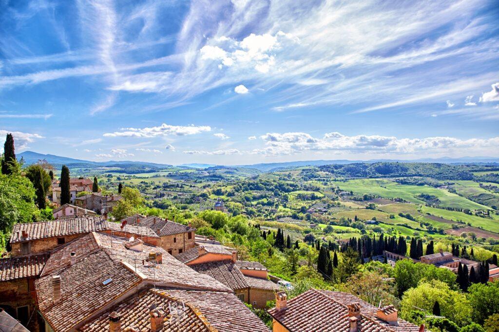 Panoramautsikt over Montepulciano i Toscana, Italia, på en vakker solrik dag. I forgrunnen sees røde tak av historiske bygninger, omgitt av frodig vegetasjon og høye sypresser. Utover i landskapet er det grønne daler, åser og jordbruksområder som strekker seg til horisonten. Den klare blå himmelen med spredte skyer gir bildet en følelse av dybde og ro. Vi har laget en omfattende guide til Toscana i Italia hvor vi blant annet forteller om fem populære byer som Firenze, Pisa, Siena, Montepulciano og Volterra. Vi gir deg også forslag til tre kjøreruter du kan ta for å oppleve vingårder og resten av Toscana. La deg inspirere av den italienske regionen som fikk sitt eget TV-program; «Den store matfesten - Toscana»