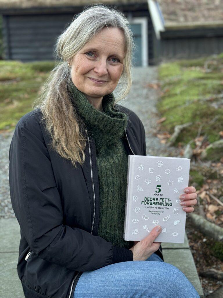 Bilde av ernæringsfysiolog Ragnhild Lekven Fimreite. Forfatteren av boken 5 trinn til bedre fettforbrenning som sitter smilende med boken sin i handen ute i naturen med trehus i bakgrunnen. Vi har fått oppskrift på sunn, hjemmelaget granola som passer perfekt til yoghurt av Ragnhild!