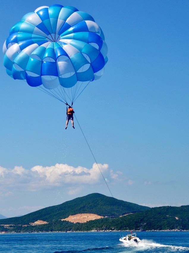 Bildet viser en person som driver med parasailing, hengende høyt oppe i luften under en stor blå og hvit fallskjerm. Personen er festet til en sele og blir dratt av en båt som kjører på vannet nedenfor. I bakgrunnen kan man se et landskap med grønne åser og fjell under en blå, skyfri himmel. Her får du ti ting du kan gjøre på Gran Canaria, Kanariøyene