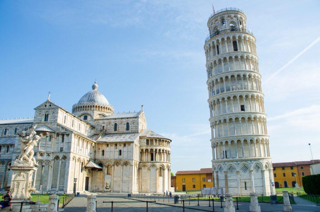 Bilde av det skjeve tårnet i Pisa, Italia, på en solrik dag med blå himmel. Til venstre i bildet er katedralen i Pisa med sin store kuppel og intrikate fasade. Foran katedralen står en statue, og til høyre sees det berømte skjeve tårnet som lener seg tydelig til siden. Rundt bygningene er det åpne områder med gress og enkelte besøkende som går rundt. Vi har laget en omfattende guide til Toscana i Italia hvor vi blant annet forteller om fem populære byer som Firenze, Pisa, Siena, Montepulciano og Volterra. Vi gir deg også forslag til tre kjøreruter du kan ta for å oppleve vingårder og resten av Toscana. La deg inspirere av den italienske regionen som fikk sitt eget TV-program; «Den store matfesten - Toscana»