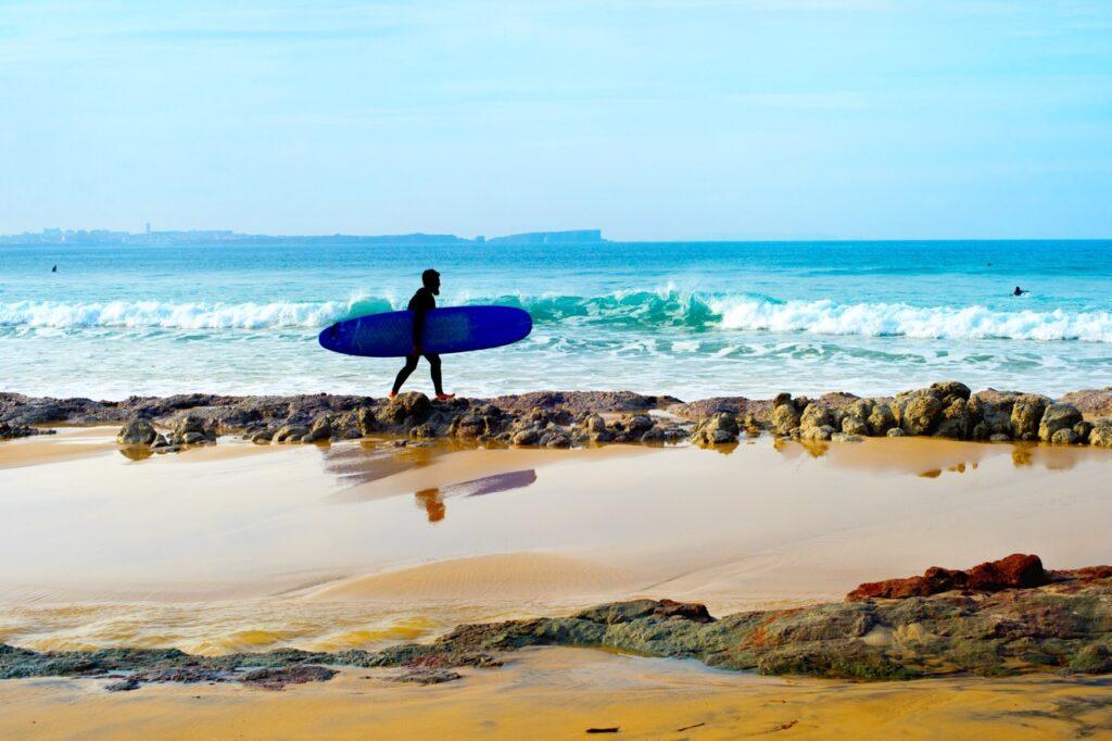 Bildet viser en surfer som går på en strand i Sagres, langs Algarve-kysten i Portugal. Surferen bærer et blått surfebrett under armen og er kledd i en våtdrakt. I bakgrunnen ruller bølgene inn mot kysten, og den klare, blå himmelen møter horisonten. Man kan også skimte en annen surfer i vannet. Vi hjelper deg med å planlegge ferien til Algarve-kysten i Portugal. Vi gir deg alt fra tips til byer; Lagos, Faro, Tavira, Albufeira og Sagres. Samt hvor du kan finne hotell langs Algarve-kysten og shoppingmulighetene.