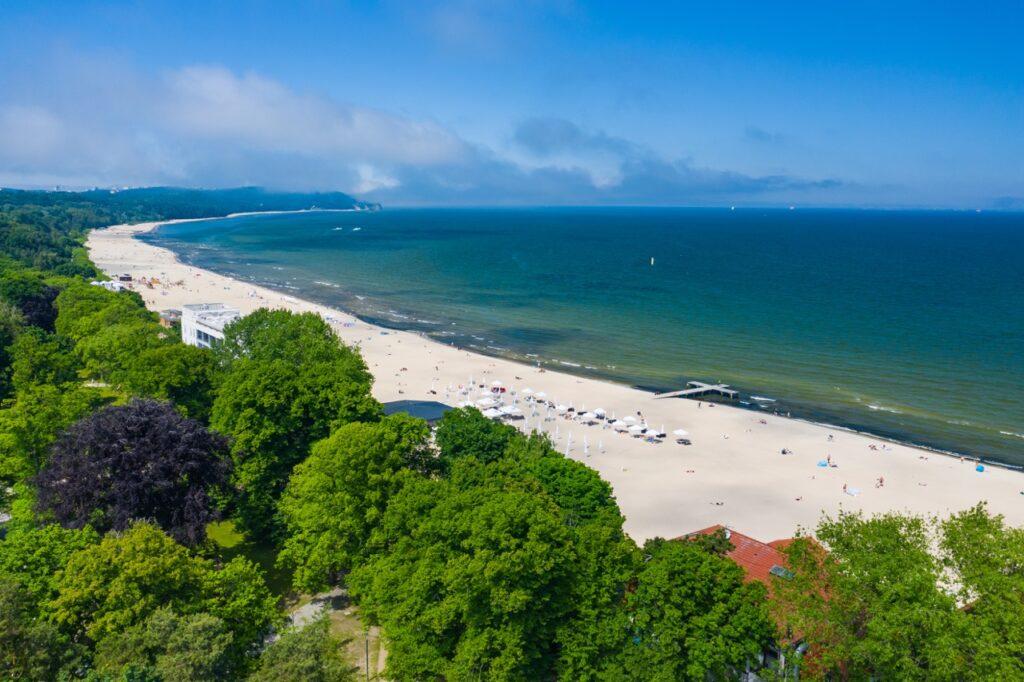 Bilde av den lange flotte stranden i Sopot, Polen. Stranden strekker seg så langt øyet kan se. Til venstre i bildet er det mange grønne frodige trær, mens til høyre i bildet ser du blå sjø. Sopot i Polen er perfekt for deg som liker å bade eller å nyte luksus på ett av spaene. Sopot har flere hotell som passer forskjellige budsjetter enten du er ute etter luksus eller budsjettvennlige hotell i Sopot. Her finner du billig ferie til sol og varme.