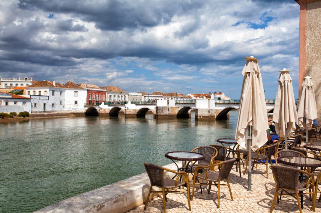 Bildet viser byen Tavira langs Algarve-kysten i Portugal. I forgrunnen er det et utendørs kaféområde med tomme bord og stoler, samt sammenfoldede parasoller. I bakgrunnen ser vi en gammel steinbro med flere buer som strekker seg over en rolig elv. På den andre siden av elven står sjarmerende, fargerike bygninger med tradisjonell portugisisk arkitektur under en dramatisk, skyet himmel. Vi hjelper deg med å planlegge ferien til Algarve-kysten i Portugal. Vi gir deg alt fra tips til byer; Lagos, Faro, Tavira, Albufeira og Sagres. Samt hvor du kan finne hotell langs Algarve-kysten og shoppingmulighetene.