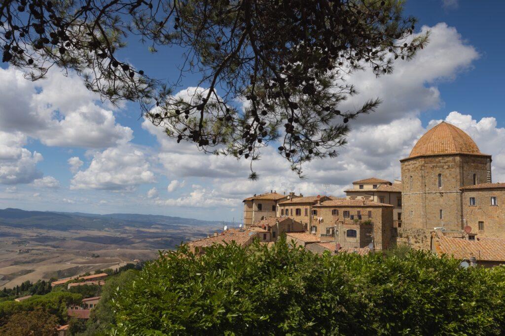 Bilde av byen Volterra i Toscana, Italia, på en dag med blå himmel og spredte skyer. Byen har historiske bygninger med røde tak, inkludert en stor rund kuppel til høyre i bildet. I forgrunnen er det frodige trær som rammer inn scenen, og utsikten strekker seg utover det bølgende toskanske landskapet med åser og daler i bakgrunnen. Vi har laget en omfattende guide til Toscana i Italia hvor vi blant annet forteller om fem populære byer som Firenze, Pisa, Siena, Montepulciano og Volterra. Vi gir deg også forslag til tre kjøreruter du kan ta for å oppleve vingårder og resten av Toscana. La deg inspirere av den italienske regionen som fikk sitt eget TV-program; «Den store matfesten - Toscana»