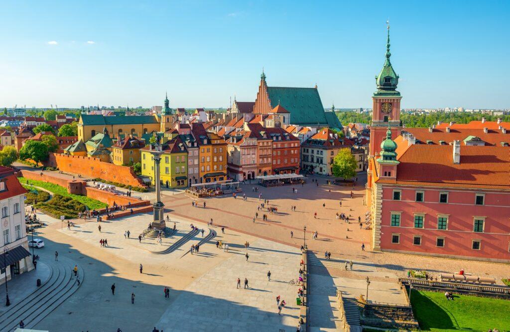 Dette bildet viser et fugleperspektiv av Slottsplassen i gamlebyen i Warszawa, Polen. I midten av plassen står Sigismund-søylen, en høy søyle med en statue på toppen. Rundt plassen er det fargerike historiske bygninger i gule, grønne og røde toner med røde tak. Til høyre ser vi det kongelige slottet med sin karakteristiske klokketårn. I bakgrunnen er en stor kirke med et grønt tak synlig. Mange mennesker går rundt på plassen og nyter det fine været. Vi gir deg tips til hva du kan se og gjøre i storbyen Warszawa. Blant annet Stare Miasto (Gamlebyen), Krakowskie Przedmieście, Łazienki-parken, Nowy Świat-gaten og Wilanów-palasset. Byen er perfekt for en storby-weekend i Europa.