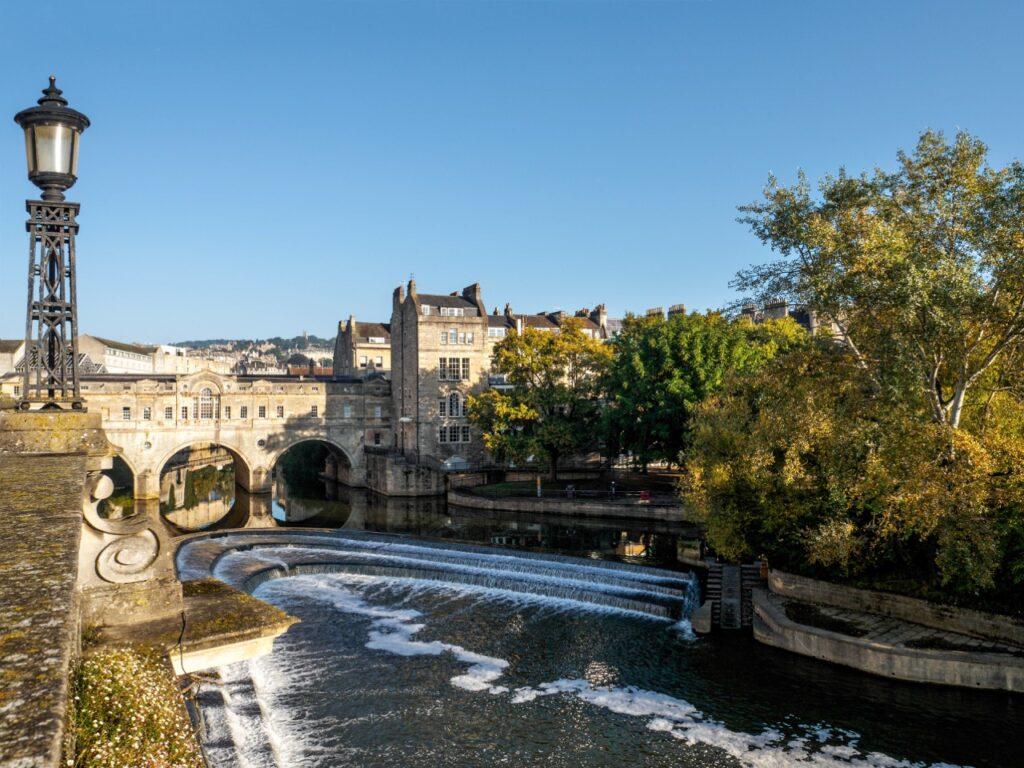 En vakker utsikt over elven i Bath, England, med en elegant steinbro i bakgrunnen. Vannet renner ned over en liten demning, og gamle bygninger og trær står langs elvebredden under en klar, blå himmel. Vi har samlet fem flotte dagsturer fra storbyen London i England, her finner du inspirasjon til turer til blant annet Oxford, Bath, Brighton, Stonehenge og Windsor Castle.