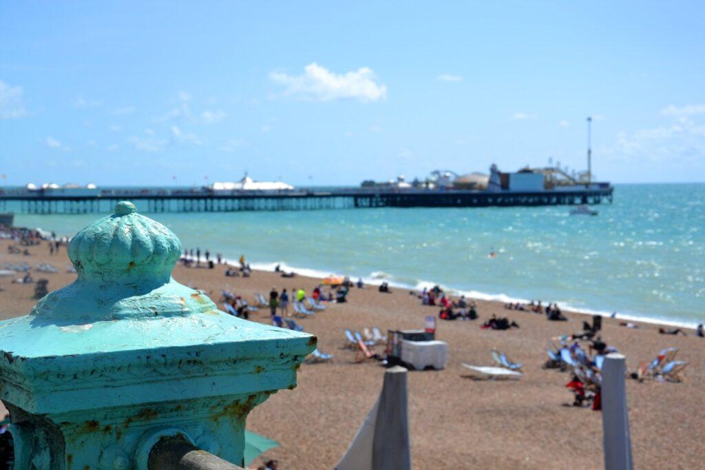 En utsikt over stranden i Brighton, England. I forgrunnen er en gammel, rustikk blågrønn rekkverkspost, og i bakgrunnen sees Brighton Pier som strekker seg ut i det blå havet. Mange mennesker soler seg og nyter dagen på stranden under en klar, blå himmel med noen få skyer. Vi har samlet fem flotte dagsturer fra storbyen London i England, her finner du inspirasjon til turer til blant annet Oxford, Bath, Brighton, Stonehenge og Windsor Castle.
