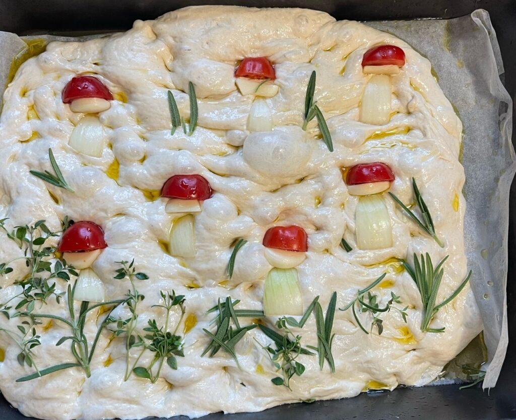 Focaccia-deig dekorert med tomater, løk, hvitløk og urter slik at det ser ut som fluesopp i et landskap.