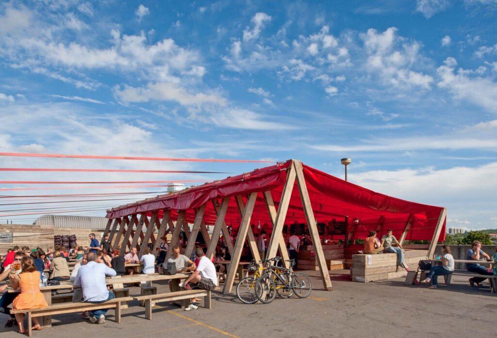 En uformell takterrasse på Frank's Cafe i London med trebenker og bord under en stor rød markise. Gjester nyter mat og drikke mens de ser ut over byens skyline på en solrik dag. Her får du syv populære takterrasser i London, England.