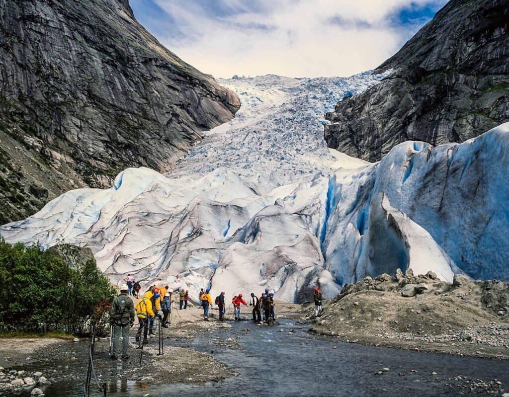 En gruppe turgåere med fargerike klær og turstaver nærmer seg en stor isbre som strekker seg oppover fjellsiden. Isbreen er blåhvit og sprekker opp i store isblokker. Området rundt isbreen er preget av steinete terreng og bratte klipper. Himmelen er delvis skyet, og fjellene rundt gir et majestetisk og dramatisk landskap. Her får du tips til syv vakre bilturer i Norge som er perfekt for norgesferien. Geiranger og Trollfjorden, Atlanterhavsveien, Lofoten, Hardanger, Rondane, Gamle Strynefjellsvegen, Senja.