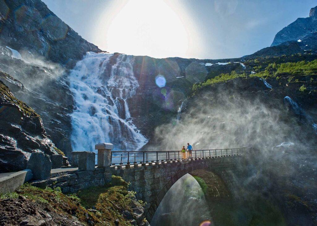 En stor foss stuper nedover fjellsiden ved Trollstigen, mens solen skinner klart i bakgrunnen. Vannet spruter og lager en fin tåke som omgir området. En steinbro med rekkverk krysser fossen, og på broen står to personer som ser ut til å nyte synet av fossen. Fjellene rundt er dekket med noe vegetasjon og snøflekker. Her får du tips til syv vakre bilturer i Norge som er perfekt for norgesferien. Geiranger og Trollfjorden, Atlanterhavsveien, Lofoten, Hardanger, Rondane, Gamle Strynefjellsvegen, Senja.