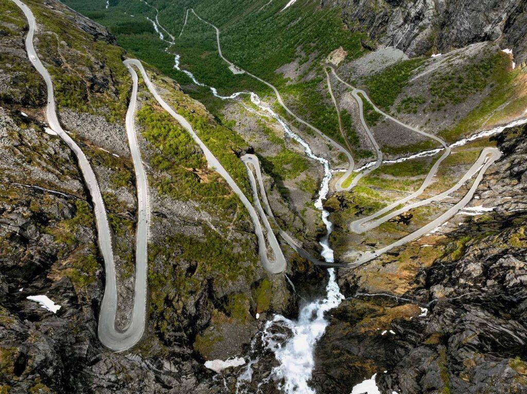 n imponerende luftfoto av Trollstigen, en bratt og svingete vei som slynger seg nedover et fjelllandskap. Veien består av mange skarpe hårnålssvinger og strekker seg over et grønt og steinete terreng. En foss kan sees i midten av bildet, der vannet strømmer nedover fjellsiden. Området er omgitt av frodig vegetasjon og bratte fjell. Her får du tips til syv vakre bilturer i Norge som er perfekt for norgesferien. Geiranger og Trollfjorden, Atlanterhavsveien, Lofoten, Hardanger, Rondane, Gamle Strynefjellsvegen, Senja.