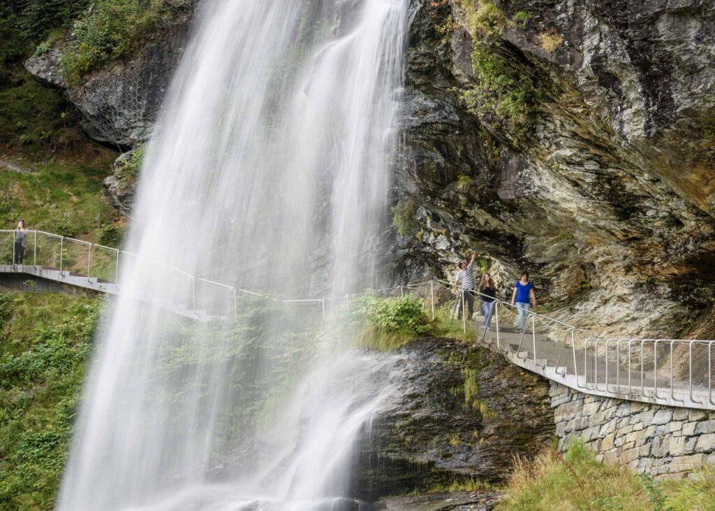 En gruppe mennesker går på en gangbro som passerer tett ved Steinsdalsfossen. Fossen kaster vann nedover klippen, og vegetasjonen rundt er grønn og frodig. Gangbroen er bygget av metall og stein, og strekker seg langs fjellveggen. Personene på broen ser ut til å nyte utsikten og omgivelsene. Her får du tips til syv vakre bilturer i Norge som er perfekt for norgesferien. Geiranger og Trollfjorden, Atlanterhavsveien, Lofoten, Hardanger, Rondane, Gamle Strynefjellsvegen, Senja.