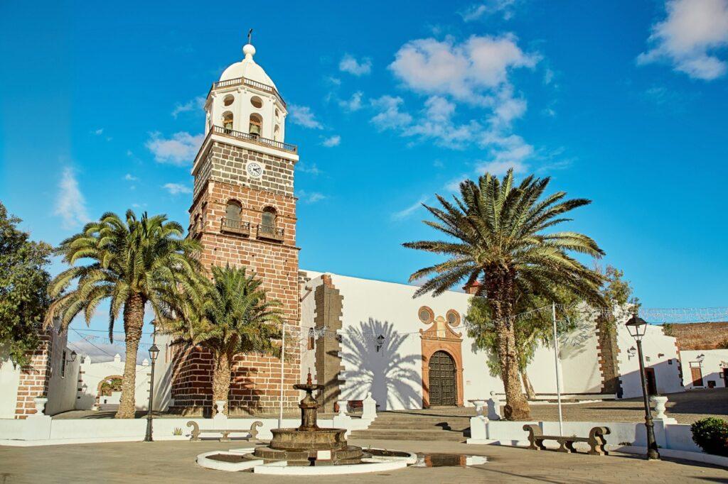 Et historisk klokketårn omgitt av palmetrær i en pittoresk by på Lanzarote, Kanariøyene. Klokketårnet står på en liten plass med en fontene i forgrunnen, omgitt av hvitmalte bygninger med tradisjonell arkitektur under en lys blå himmel.