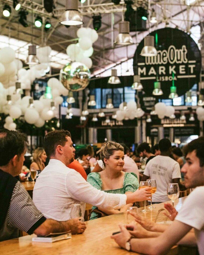 Bildet viser et matmarked, spesifikt Time Out Market i Lisboa. Folk sitter ved lange trebord og nyter mat og drikke. En mann i en hvit skjorte og en kvinne i en grønn kjole sitter sammen og skåler med glassene sine. Rundt dem er det andre besøkende som også nyter måltidene sine. Taket er dekorert med hvite ballonger og flere hengende lamper, og en stor diskokule henger i midten, som reflekterer lysene i rommet. Atmosfæren virker livlig og sosial, med mange mennesker samlet for å nyte markedets tilbud. Her finner du tips til ni matmarkeder i populære europeiske storbyer som Paris, Berlin, Roma, Barcelona, København, Lisboa og London.