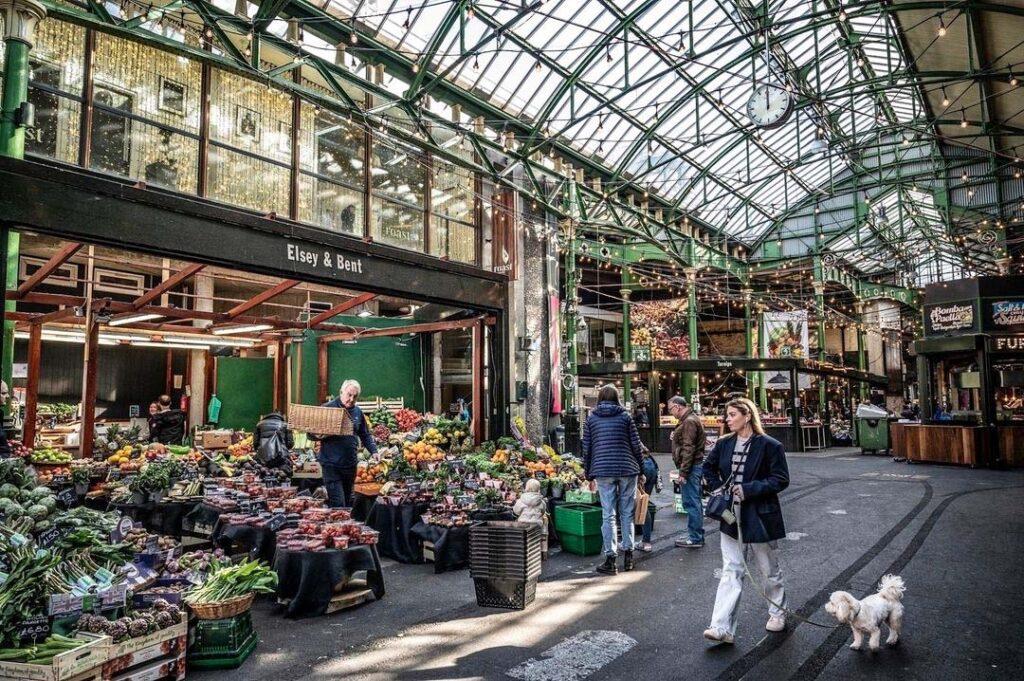 Bildet viser et travelt innendørs matmarked i London, nærmere bestemt Borough Market. Flere mennesker går rundt og handler frukt og grønnsaker fra forskjellige boder. En eldre mann bærer en kasse med varer gjennom markedet. Markedet har et høyt tak med grønne metallbjelker og glasspaneler som slipper inn naturlig lys. Lyslenker og dekorative lamper henger ned fra taket, og gir en varm og innbydende atmosfære. Her finner du tips til ni matmarkeder i populære europeiske storbyer som Paris, Berlin, Roma, Barcelona, København, Lisboa og London.