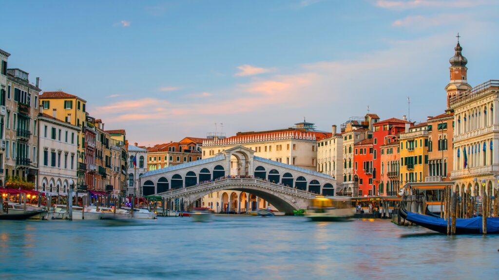 Bildet viser den berømte Rialto-broen i Venezia, Italia, som strekker seg over Canal Grande. Broen er et av Venezias mest ikoniske landemerker, og den er omgitt av fargerike bygninger med typisk venetiansk arkitektur. I forgrunnen ser man flere båter og gondoler som glir langs kanalen, noe som gir en følelse av liv og bevegelse. På broen og langs kanalens bredder er det mennesker som nyter den vakre utsikten. Her får du en guide til hav du bør se og gjøre i Venezia, Italia. Vi anbefaler blant annet å ta en gondoltur, Besøk Markusplassen (Piazza San Marco), Utforsk Rialto-broen og markedet, Oppdag øyene Murano, Burano og Torcello og Besøk Dogepalasset (Palazzo Ducale.