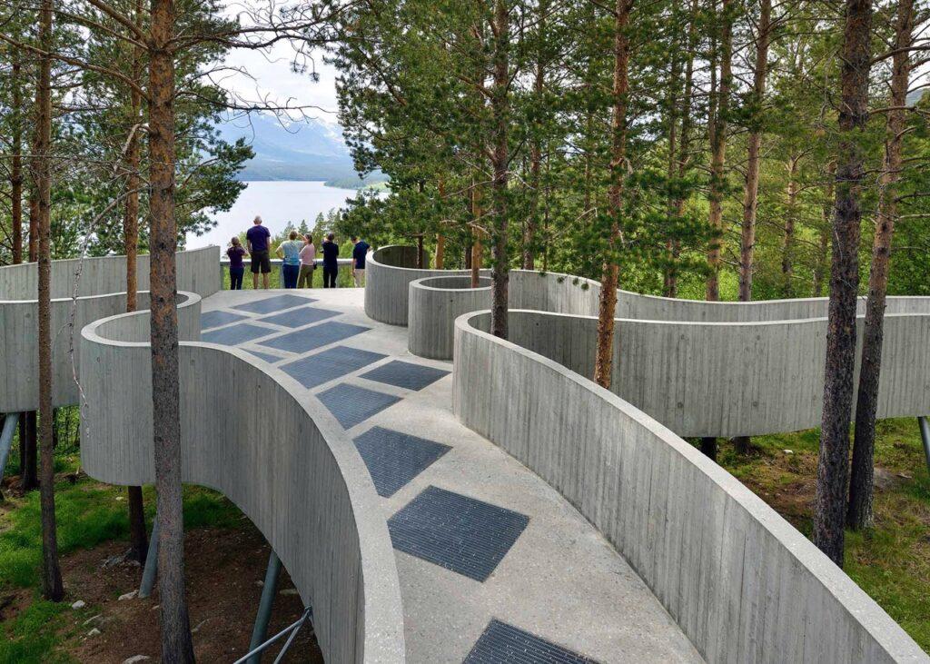 En moderne utsiktsplattform laget av betong, som slynger seg gjennom en skog av furutrær. Flere personer står på enden av plattformen og ser ut over en innsjø og fjellene i bakgrunnen. Plattformen har buede former og skråstilte flater som gir et kunstnerisk preg. Trærne rundt plattformen gir skygge og rammer inn utsikten mot det vakre landskapet. Her får du tips til syv vakre bilturer i Norge som er perfekt for norgesferien. Geiranger og Trollfjorden, Atlanterhavsveien, Lofoten, Hardanger, Rondane, Gamle Strynefjellsvegen, Senja.