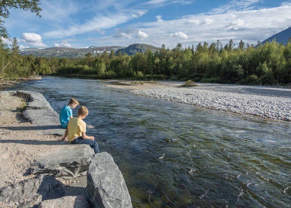 To barn sitter på en steinkant ved siden av en elv i Rondane. De ser ut til å leke med vannet, og en av dem har føttene i elven. Landskapet rundt elven er frodig med trær, og i bakgrunnen kan man se fjell med snøflekker under en klar, blå himmel med spredte skyer. Elven flyter rolig gjennom det naturskjønne området. Her får du tips til syv vakre bilturer i Norge som er perfekt for norgesferien. Geiranger og Trollfjorden, Atlanterhavsveien, Lofoten, Hardanger, Rondane, Gamle Strynefjellsvegen, Senja.