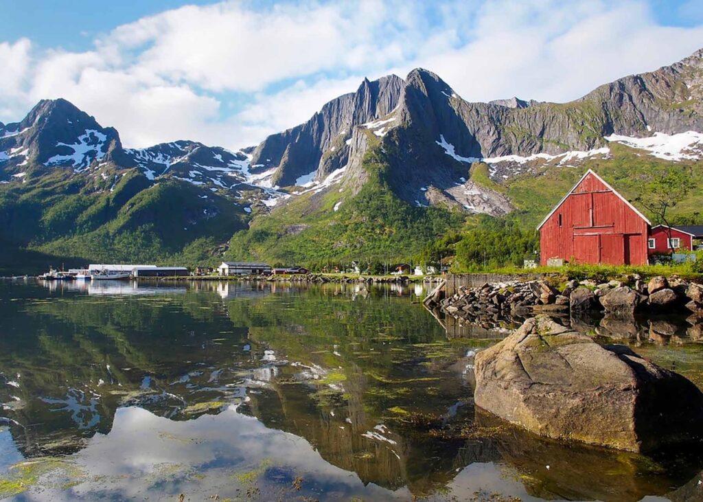 En pittoresk fjordscene på Senja med en rød låve ved vannkanten. Bakgrunnen domineres av majestetiske, snødekte fjell som speiler seg i det stille vannet. Noen små bygninger og en brygge med båter kan også sees langs vannkanten. Himmelen er blå med noen spredte skyer, og vegetasjonen rundt er frodig og grønn. Her får du tips til syv vakre bilturer i Norge som er perfekt for norgesferien. Geiranger og Trollfjorden, Atlanterhavsveien, Lofoten, Hardanger, Rondane, Gamle Strynefjellsvegen, Senja.