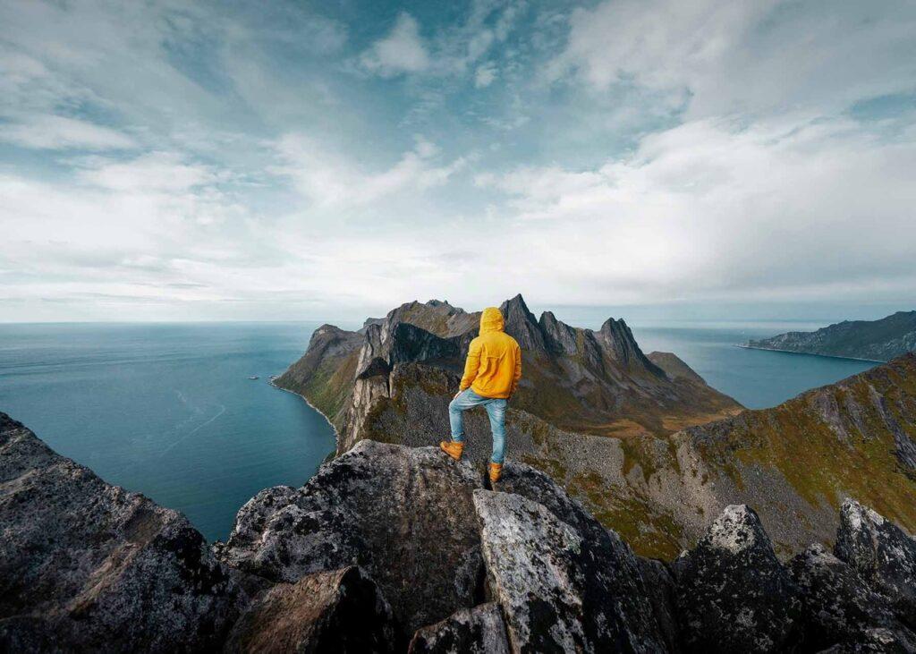 En person iført en gul jakke og jeans står på en fjellknaus og ser utover det dramatiske landskapet på Senja. Foran personen strekker fjellene seg ut mot havet med bratte klipper og spisse topper. Under ligger det blå havet, og horisonten er dekket av en lett skyet himmel. Utsikten gir et imponerende og majestetisk inntrykk av naturen. Her får du tips til syv vakre bilturer i Norge som er perfekt for norgesferien. Geiranger og Trollfjorden, Atlanterhavsveien, Lofoten, Hardanger, Rondane, Gamle Strynefjellsvegen, Senja.