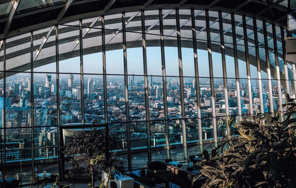 Utsikt over Londons skyline gjennom store, buede glassvinduer fra Sky Garden takterrasse. Innendørs plantepynt og sittegrupper kan sees i forgrunnen. Her får du syv populære takterrasser i London, England.