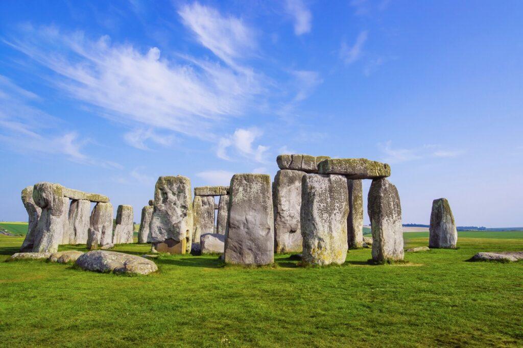 Et bilde av Stonehenge, den berømte forhistoriske steinformasjonen i England. De massive, værslitte steinene står på en grønn eng under en klar, blå himmel med lette, hvite skyer som strekker seg over horisonten. Vi har samlet fem flotte dagsturer fra storbyen London i England, her finner du inspirasjon til turer til blant annet Oxford, Bath, Brighton, Stonehenge og Windsor Castle.