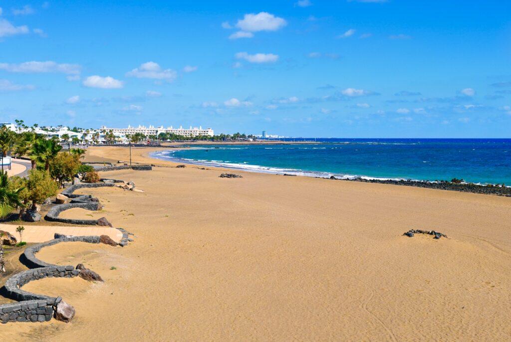 En vidstrakt strand på Lanzarote, Kanariøyene, med gyllen sand og det blå havet som strekker seg mot horisonten. Langs stranden er det promenader med palmer og frodige planter, og i bakgrunnen kan man se hvite bygninger og hotellkomplekser under en klar blå himmel.