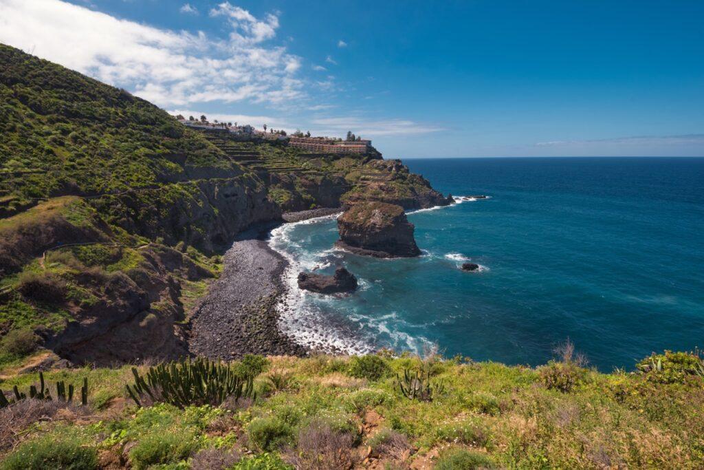 En steinete kystlinje på Tenerife, Kanariøyene, med bratte grønne klipper som stuper ned i det klare blå havet. Langs kysten er det noen små steinstrender, og vegetasjonen er frodig og grønn. Tenerife er den største Kanariøyen og har Spanias høyeste fjell, Teide. Hovedstaden er Santa Cruz de Tenerife. Her kan du finne billige reiser til Tenerife, samt hotell på den populære kanariøyen.