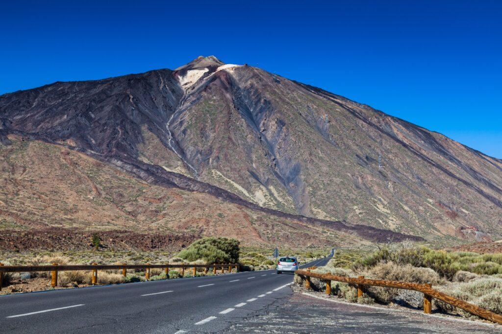 En bil kjører langs en vei som leder mot fjellet Teide på Tenerife, Kanariøyene. Fjellet er dekket av mørke vulkanske steiner og vegetasjon, og den klare blå himmelen gir en skarp kontrast til landskapet. Veien er omgitt av busker og små trær. Tenerife er den største Kanariøyen og har Spanias høyeste fjell, Teide. Hovedstaden er Santa Cruz de Tenerife. Her kan du finne billige reiser til Tenerife, samt hotell på den populære kanariøyen.