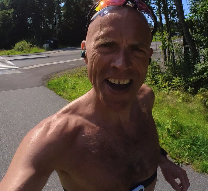 Bildet viser løpeeksperten Andreas Gossner fra løpetrening.no som løper utendørs på en solrik dag. Han har på seg en pulsmåler over bryster og solbriller fra Oakley til trening. På bildet ser vi mannen smilende og løpende på en asfaltert vei omgitt av grønn natur og trær mens han tar en selfie. Dette bildet formidler glede ved løping og en aktiv livsstil utendørs i vakre omgivelser. Løpeeksperten Andreas Gossner fra løpetrening.no gir deg et treningsprogram for halvmaraton som gjør at du kan løpe ditt raskeste halvmaraton noen sinne!