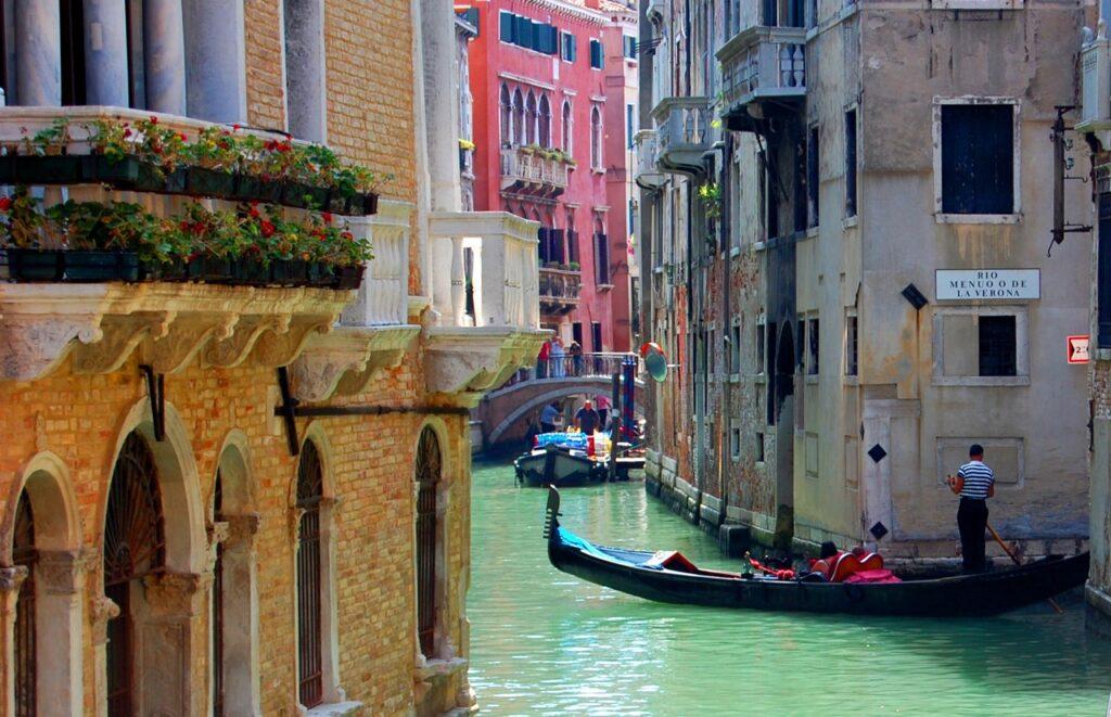 Bildet viser en typisk kanal i Venezia, Italia, med gondoler som seiler gjennom det smaragdgrønne vannet. På venstre side er det bygninger med fasader av gul murstein, dekorert med balkonger fylt med blomsterkasser. På høyre side er det en smal kanal med flere vinduer og en skilt som viser "Rio Menuo de la Verona". En gondolier, kledd i en stripete skjorte, styrer gondolen langs den smale kanalen. I bakgrunnen er det flere vakre bygninger i venetiansk stil og en liten bro som forbinder de to sidene av kanalen. Her får du en guide til hav du bør se og gjøre i Venezia, Italia. Vi anbefaler blant annet å ta en gondoltur, Besøk Markusplassen (Piazza San Marco), Utforsk Rialto-broen og markedet, Oppdag øyene Murano, Burano og Torcello og Besøk Dogepalasset (Palazzo Ducale.