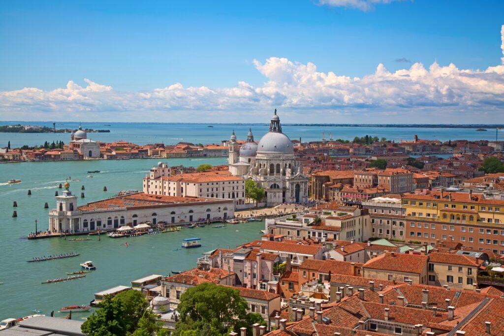 Bilde av Venezia, Italia, tatt fra en høy vinkel som gir utsikt over den vakre lagunen og byens ikoniske arkitektur. I forgrunnen ser man flere bygninger med røde takstein og en stor hvit kuppel, som sannsynligvis er Basilica di Santa Maria della Salute. Flere båter og gondoler beveger seg på det blågrønne vannet i kanalene. I bakgrunnen strekker den blå himmelen seg over havet med noen få hvite skyer. Bildet fanger essensen av Venezias unike kombinasjon av historisk arkitektur og pittoreske vannveier. Her får du en guide til hav du bør se og gjøre i Venezia, Italia. Vi anbefaler blant annet å ta en gondoltur, Besøk Markusplassen (Piazza San Marco), Utforsk Rialto-broen og markedet, Oppdag øyene Murano, Burano og Torcello og Besøk Dogepalasset (Palazzo Ducale.