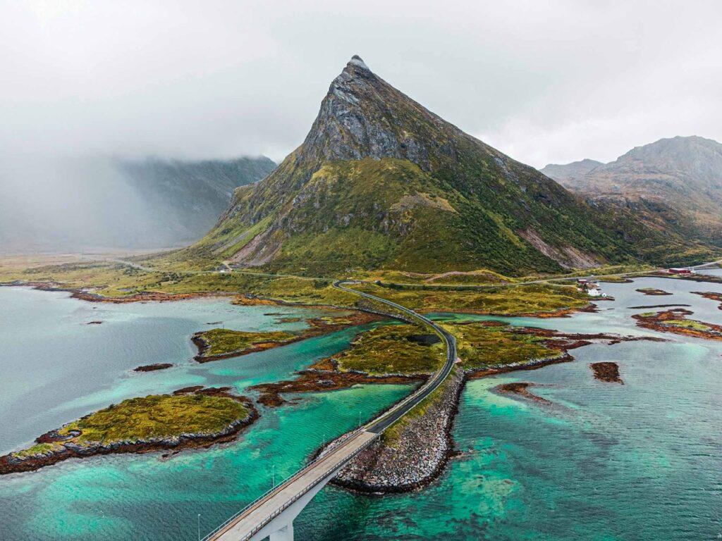 Et luftfoto som viser en lang bro i Lofoten som strekker seg over klart, turkisfarget vann og forbinder små øyer. Den spisse toppen av Volandstinden stiger majestetisk opp fra det grønne landskapet, omgitt av lavtliggende skyer og tåkedekte fjell i bakgrunnen. Her får du tips til syv vakre bilturer i Norge som er perfekt for norgesferien. Geiranger og Trollfjorden, Atlanterhavsveien, Lofoten, Hardanger, Rondane, Gamle Strynefjellsvegen, Senja.