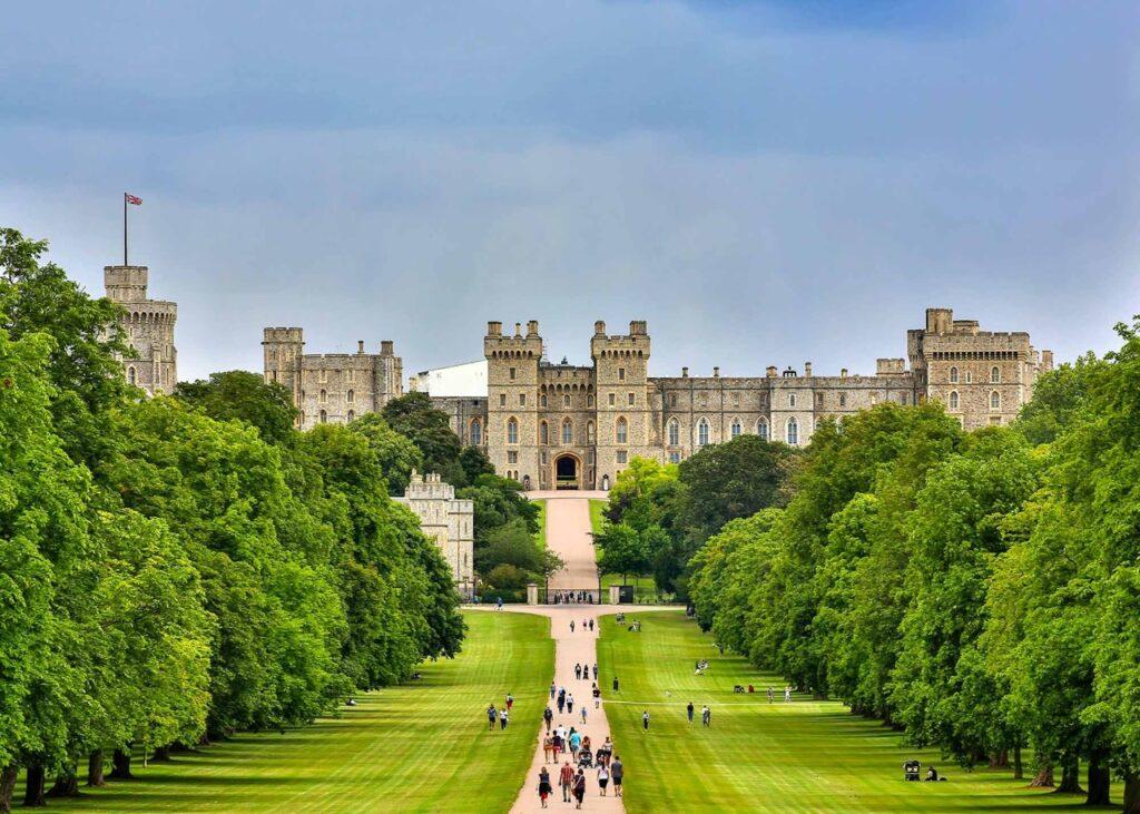 Et bilde av Windsor Castle i England, sett fra en avstand langs en bred gressdekket vei omgitt av trær. Slottet med sine imponerende tårn og steinvegger ruver majestetisk under en overskyet himmel, mens besøkende spaserer opp mot bygningen. Vi har samlet fem flotte dagsturer fra storbyen London i England, her finner du inspirasjon til turer til blant annet Oxford, Bath, Brighton, Stonehenge og Windsor Castle.