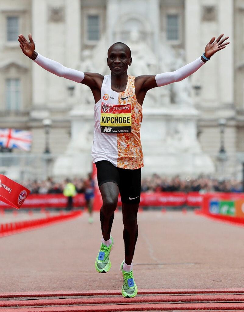 Bildet viser den kenyanske maratonløperen Eliud Kipchoge i det han krysser målstreken under London Marathon. Han er kledd i en løpetrøye med en halvt oransje og hvit design, sorte løpeshorts og lysegrønne løpesko. Kipchoge har armene løftet i triumf og smiler bredt, tydelig fornøyd med prestasjonen. I bakgrunnen er det en stor, staselig bygning og flere tilskuere som heier. Bildet fanger øyeblikket av seier og glede etter en imponerende prestasjon i maratonløpet, og illustrerer hvordan et smil kan være en naturlig reaksjon på suksess under trening og konkurranse. Eliud Kipchoge, for mange kjent som mannen med verdens raskeste, uoffisielle maraton-rekord, er kjent for å bevist benytte seg av ansiktsmimikk for å roe seg ned og holde ut når han løper.