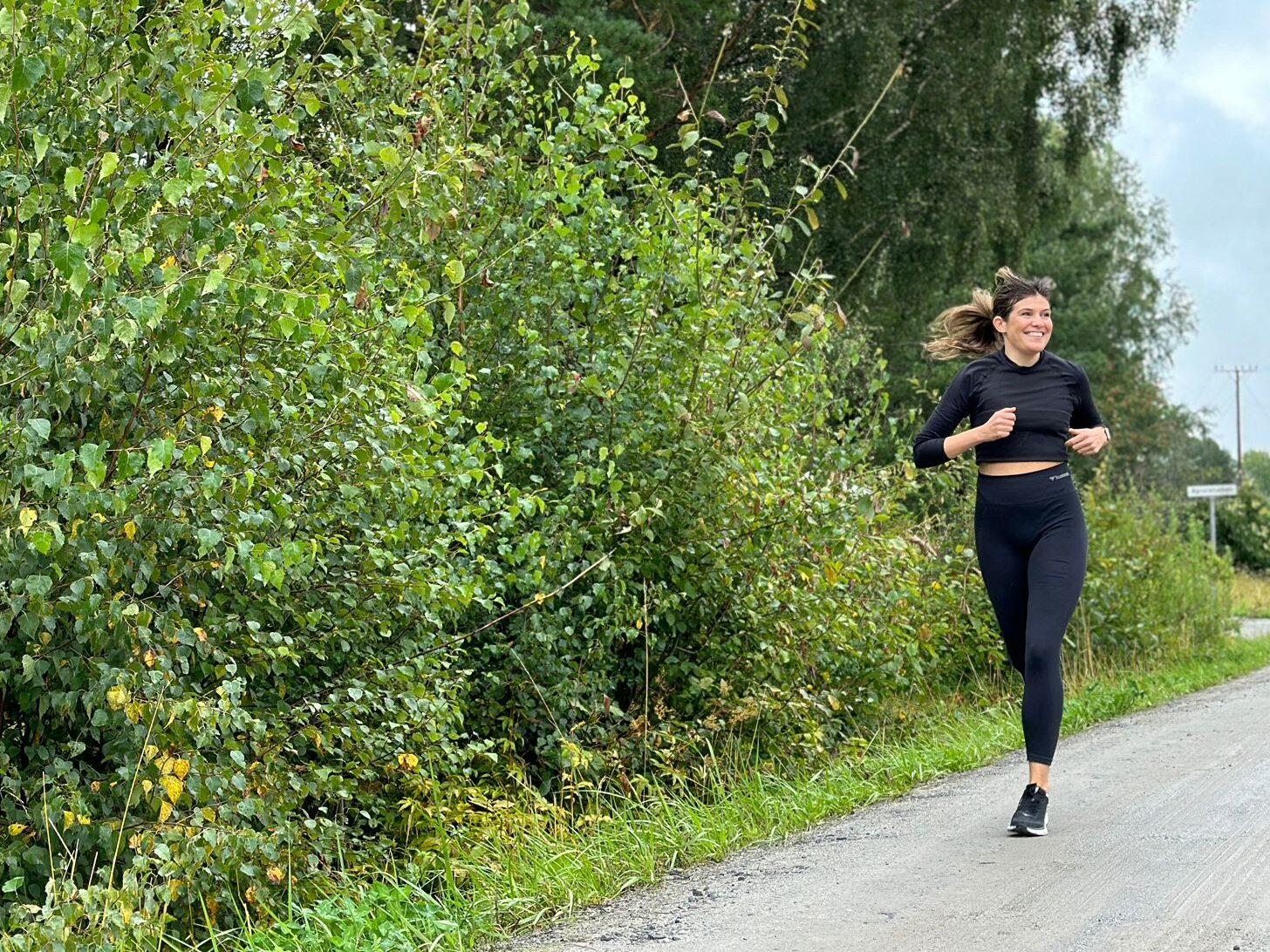 Bildet viser en kvinne som jogger langs en vei omgitt av grønn, frodig vegetasjon. Hun er kledd i svarte treningstights og en svart langermet topp, og smiler bredt mens hun løper. Hun ser avslappet og glad ut, og bildet formidler en følelse av glede og energi knyttet til løping i naturen. Bakgrunnen viser høye trær og busker, og det ser ut til å være en overskyet dag, men stemningen er likevel positiv og oppfriskende. Bildet understreker viktigheten av å smile og ha det gøy mens man trener. Vi forklarer hvorfor du bør smile når du trener, og hva som da skjer!