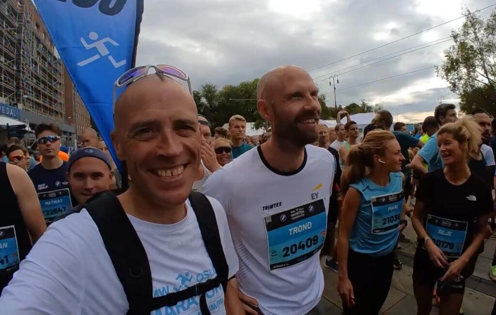 Bildet viser en gruppe mennesker som står klare ved starten av Oslo Maraton. I forgrunnen er to smilende menn kledd i løpsklær, hvorav den ene har en hvit T-skjorte med påskriften "Trond" og et startnummer på brystet. Personen ved siden av ham har solbriller på hodet og ser ut til å være en fartsholder, da det er en blå banner med et løpesymbol i bakgrunnen. Rundt dem er det flere deltakere, alle klare til å starte løpet, med forskjellige løpsnumre og T-skjorter. Stemningen virker lett og spent, med forventning om løpet som snart skal begynne. Løpetrener Andreas Gossner fra Løpetrening.no forteller hvordan du kan bruke et fremtidig maraton eller konkurranse som motivasjon og inspirasjon til treningen din!