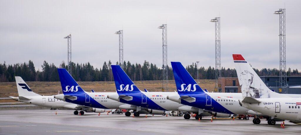 Bildet viser flere passasjerfly som står parkert på en flyplass. Flyene tilhører ulike skandinaviske flyselskaper, inkludert SAS, Finnair, og Norwegian. Det er en rekke fly som står på linje med flyenes haler synlige, alle med sine respektive logoer og farger. Området rundt flyene er åpent, med noen lyktestolper og skog i bakgrunnen. Vi forteller hva reiseforsikringen dekker ved eventuell flystreik, samt hva du kan kreve av flyselskapene ved økonomiske tap ved flystreik.