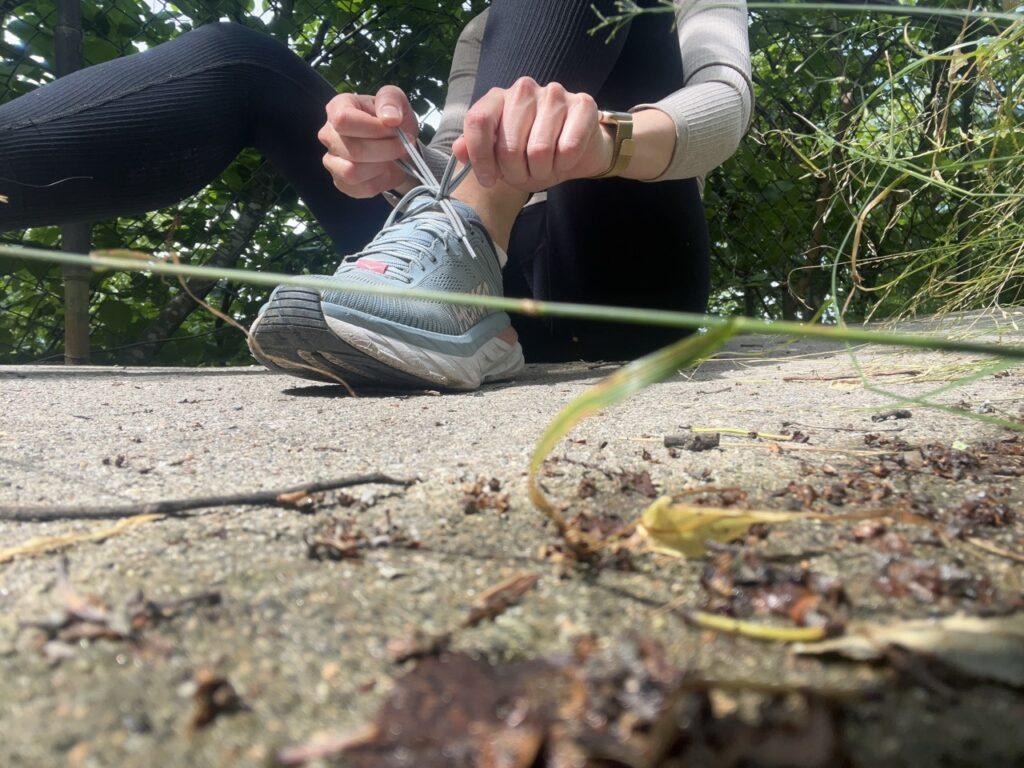 Bilde av en kvinne som sitter på bakken og knytter skoene sine. Hun har på seg en svart treningstights, en beige treningstopp fra Aimn og lyseblå løpesko fra Hoka (Hoka Clifton). På bildet ser man grønne go frodige trær og gresstrå. Løpeekspert Andreas Gossner fra Løpetrening.no svarer: Kan alle løpe et 100 miles ultraløp? Og hvordan får man det til?