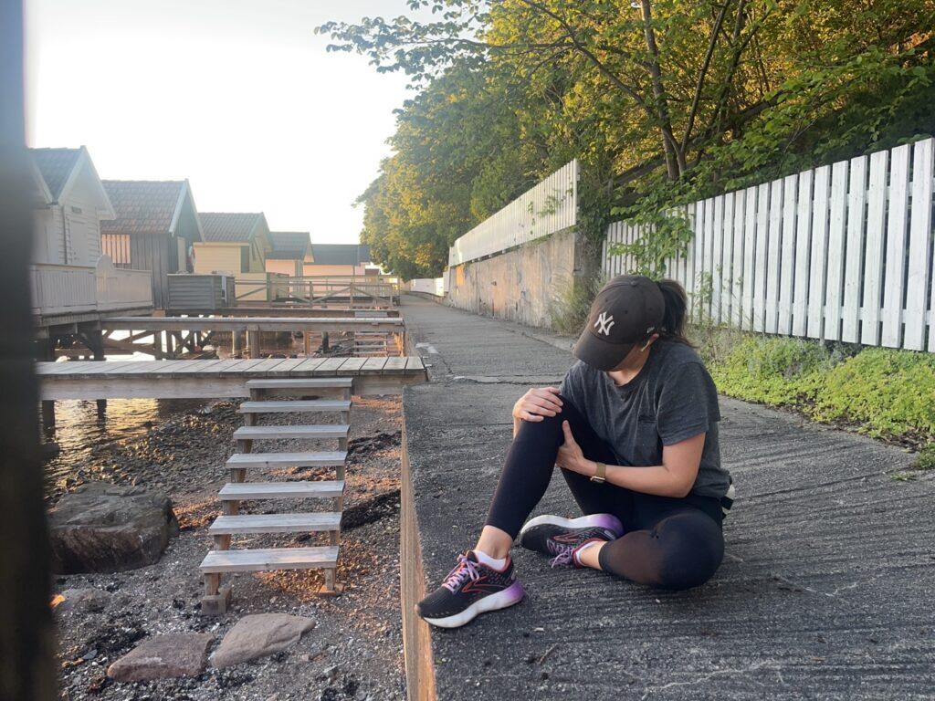 Bilde av en kvinne som sitter på en gangvei ved små badehus ved Oslofjorden. Hun har på seg en grå treningst-skjorte, en svart NY-Yankees caps og svarte tights. Hun holder på kneet sitt fordi hun kjenner smerter og vondter og har en smartklokke på håndleddet. Bakgrunnen viser små badehus og blå himmel ved Oslofjorden. Solen skinner, og det er en lys og fin dag. Løpeekspert Andreas Gossner fra Løpetrening.no forteller