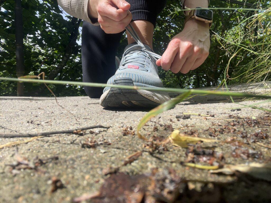 Bilde av en kvinne som sitter på bakken og knytter skoene sine. Hun har på seg en svart treningstights, en beige treningstopp fra Aimn og lyseblå løpesko fra Hoka (Hoka Clifton). På bildet ser man grønne go frodige trær og gresstrå. Løpeekspert Andreas Gossner fra Løpetrening.no svarer: Løpeeksperten svarer, Hva gjør man når man mister motivasjonen?