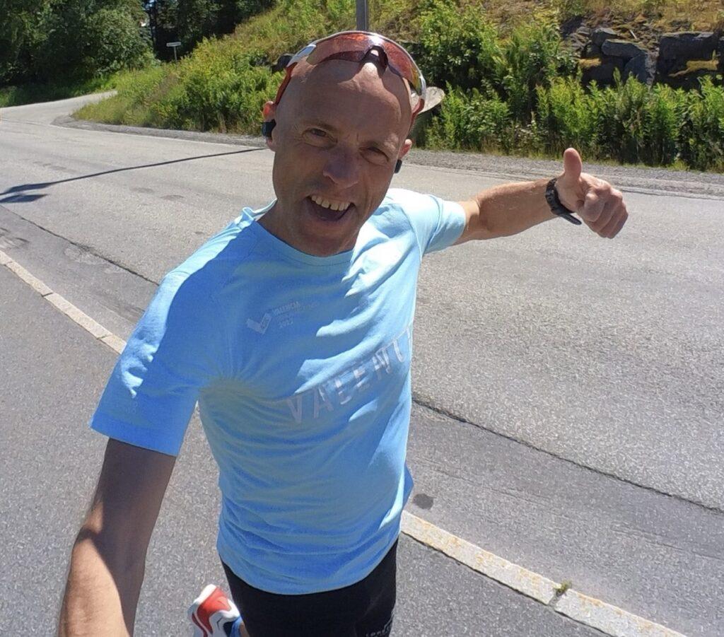 Bildet viser løpeeksperten Andreas Gossner fra løpetrening.no som løper utendørs på en solrik dag. Han har på seg en lyseblå t-skjorte og solbriller fra Oakley til trening. På bildet ser vi mannen smilende og løpende på en asfaltert vei omgitt av grønn natur og trær mens han tar en selfie. Dette bildet formidler glede ved løping og en aktiv livsstil utendørs i vakre omgivelser. Løpeeksperten Andreas Gossner fra løpetrening.no forteller hvorfor det er store fordeler med riktig oppvarming og hvordan det gjøres.