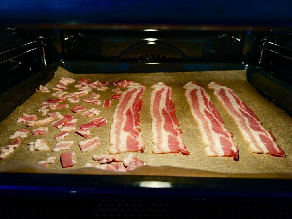 Dette bildet viser en stekeplate med bakepapir i en ovn, der det er plassert flere skiver med bacon. På venstre side av stekeplaten er det mindre biter av bacon, mens på høyre side ligger lengre, hele skiver bacon. Det ser ut som baconet er i ferd med å stekes i ovnen, kanskje for å lage sprø baconbiter til en rett, eller for å tilberede frokost