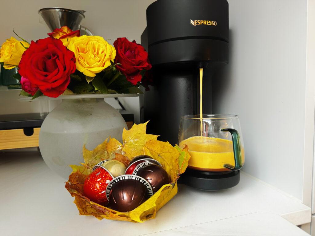 Bildet viser en Nespresso kaffemaskin i aksjon, som lager en kopp kaffe som helles i et glasskrus. Ved siden av kaffemaskinen står en vase med friske roser i røde og gule nyanser, noe som gir et livlig og fargerikt preg til scenen. Foran vasen er en liten skål laget av høstblader, som holder flere kaffekapsler i ulike farger. Denne kreative skålen tilfører et naturlig og unikt element til kaffehjørnet, som kombinerer praktisk funksjon med estetisk dekorasjon.