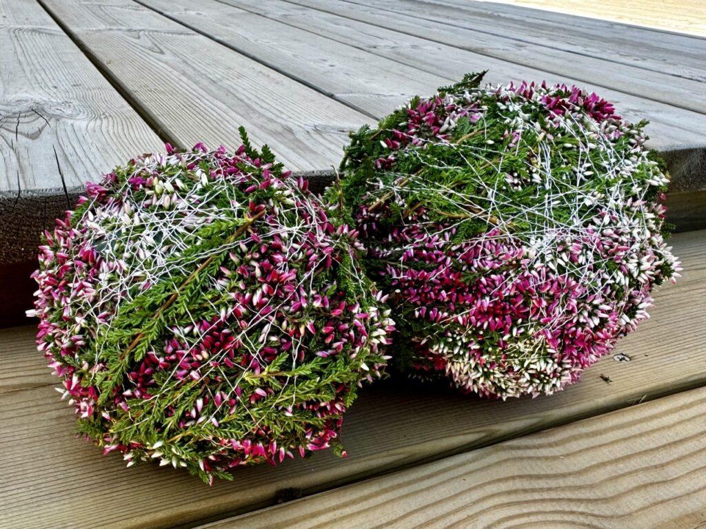 Bildet viser to håndlagde lyngballer som ligger på et trebord. Lyngballene er dekorert med et nøye arrangert mønster av tynne, hvite tråder som er surret rundt, og de er sammensatt av små blomstrende planter i nyanser av rosa, hvit og grønn. De friske fargene og de detaljerte teksturene på lyngplantene gir et naturlig og livlig preg til arrangementet. Treverket i bakgrunnen har en varm og rustikk tone som komplementerer de grønne og rosa fargene i lyngballene, noe som gir bildet en harmonisk og estetisk appell.