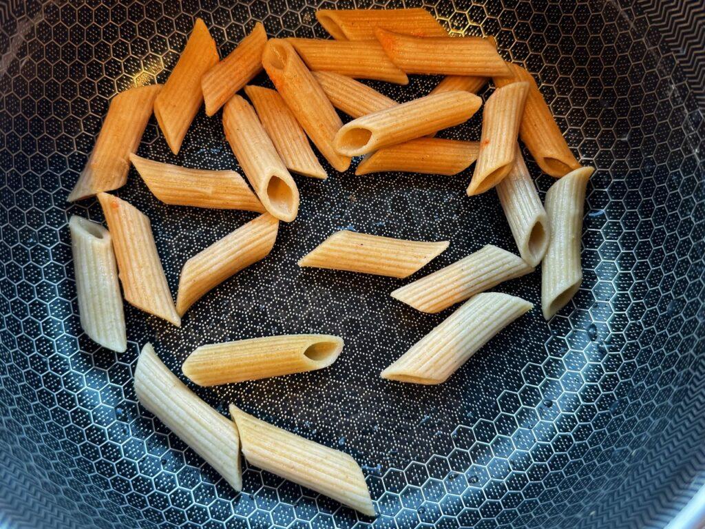 Bildet viser en kjele med en unik, geometrisk mønstret overflate, hvor flere ukokte eller lett kokte pennepastabiter ligger spredt i bunnen. Pastaen er av fullkornstypen, med en lys brunfarge, og er tilfeldig plassert på kjelens mørke overflate. Mønsteret i kjelen skaper en kontrast mot pastabitene, som er i varierende posisjoner.