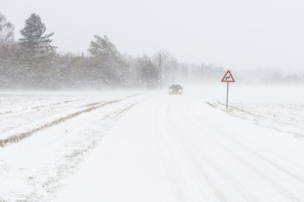 Bildet viser snødekket landevei med et trafikkskilt og en bil i det fjerne.