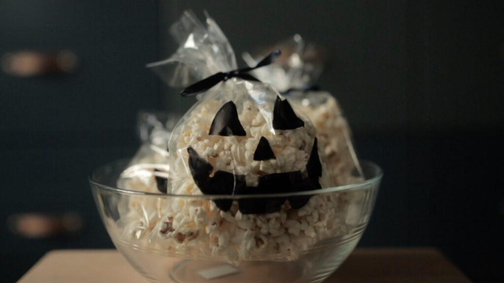 Bildet viser gjennomsiktige poser fylt med popcorn, plassert i en glassbolle. Hver pose er dekorert med sorte gresskar-ansikter laget av papir, og er bundet med svarte bånd på toppen for å gi en ekstra halloween-stemning. Posene gir inntrykk av å være små gresskar og utgjør en kreativ og morsom snacksidé til halloween-festen. Belysningen er dempet, noe som gir bildet et koselig og litt mystisk preg.